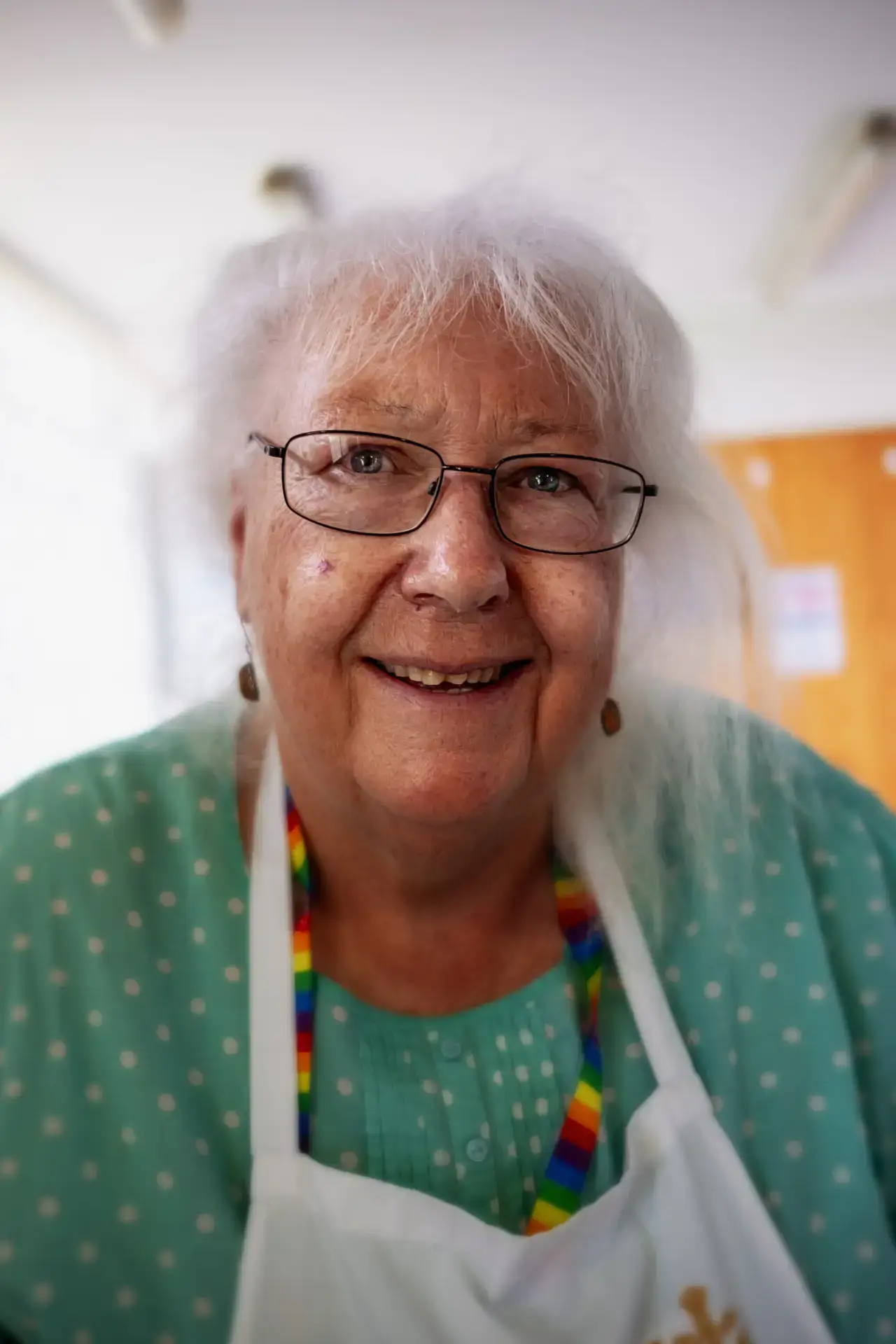 Colour portrait of an older woman with white hair and glasses, wearing a rainbow lanyard, apron, and green polka dot blouse, smiling warmly indoors.