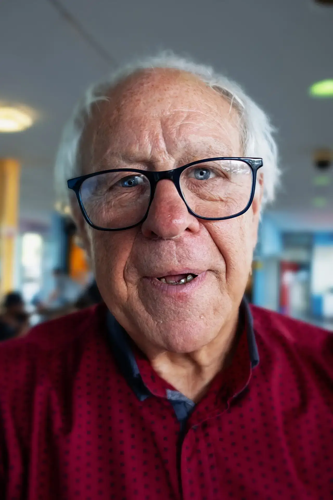 Elderly man with glasses wearing a red patterned shirt indoors.