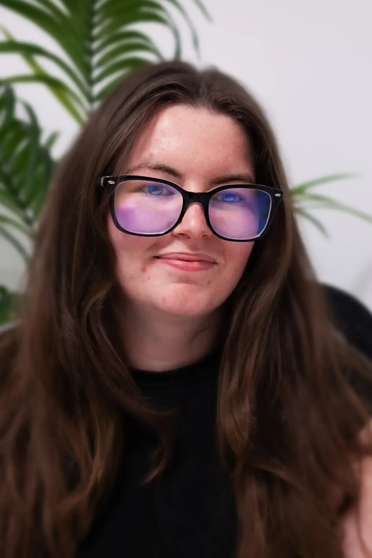 Young woman with long brown hair and glasses smiling indoors.