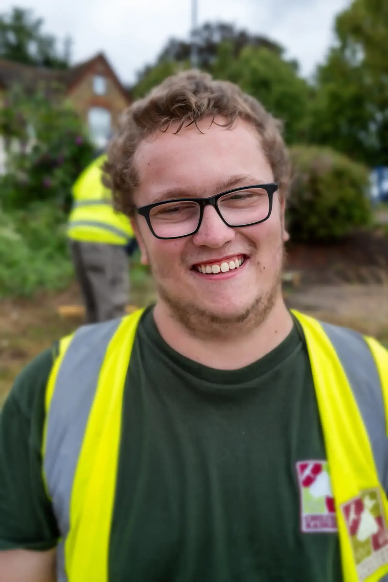 Young man in glasses and high-visibility vest smiling outdoors.