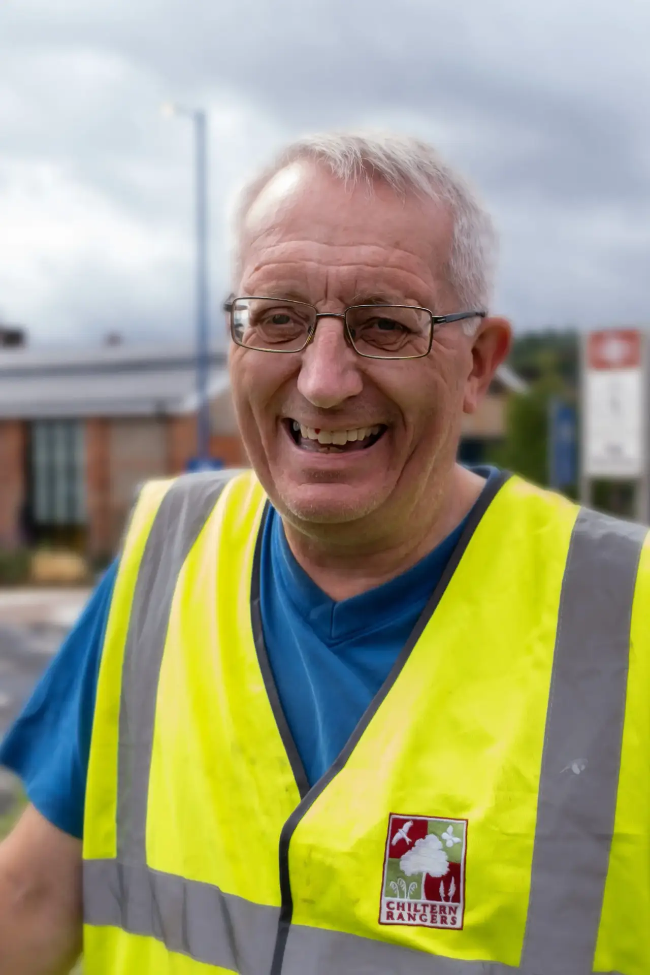 Older man in glasses wearing a high-visibility vest and smiling outdoors.