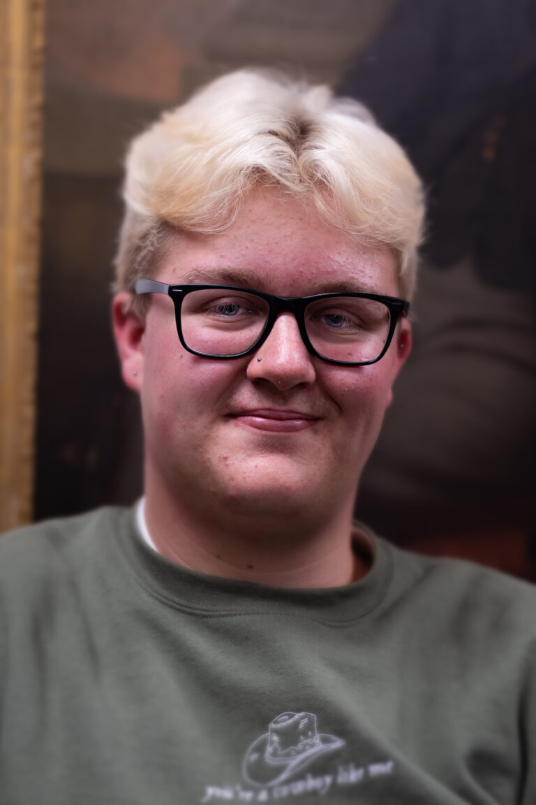 Young person with blonde hair, glasses, and nose stud wearing a green sweatshirt