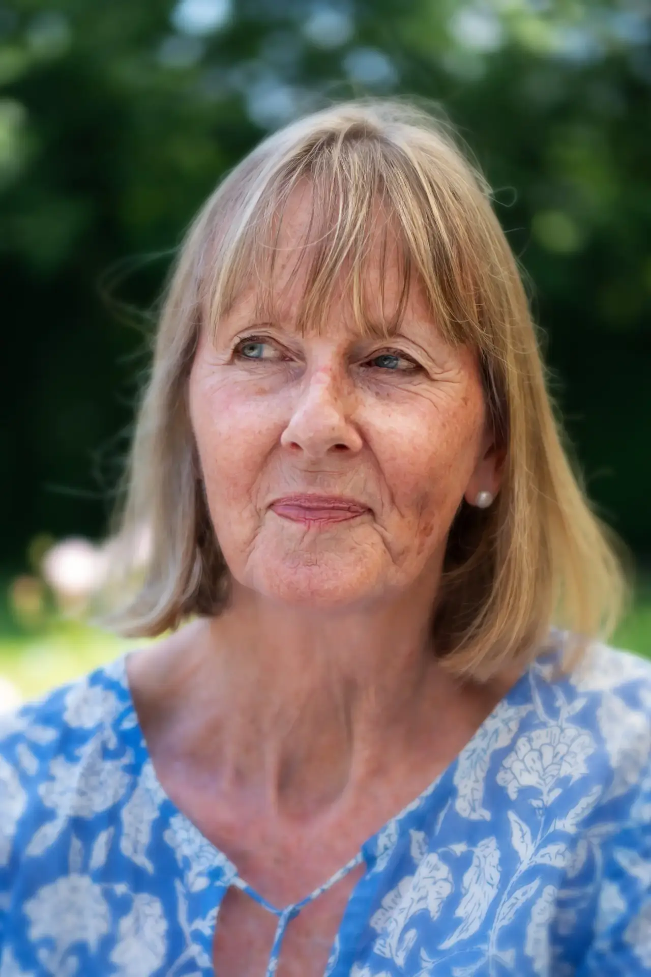 Older woman in a blue patterned blouse looking thoughtful outdoors.