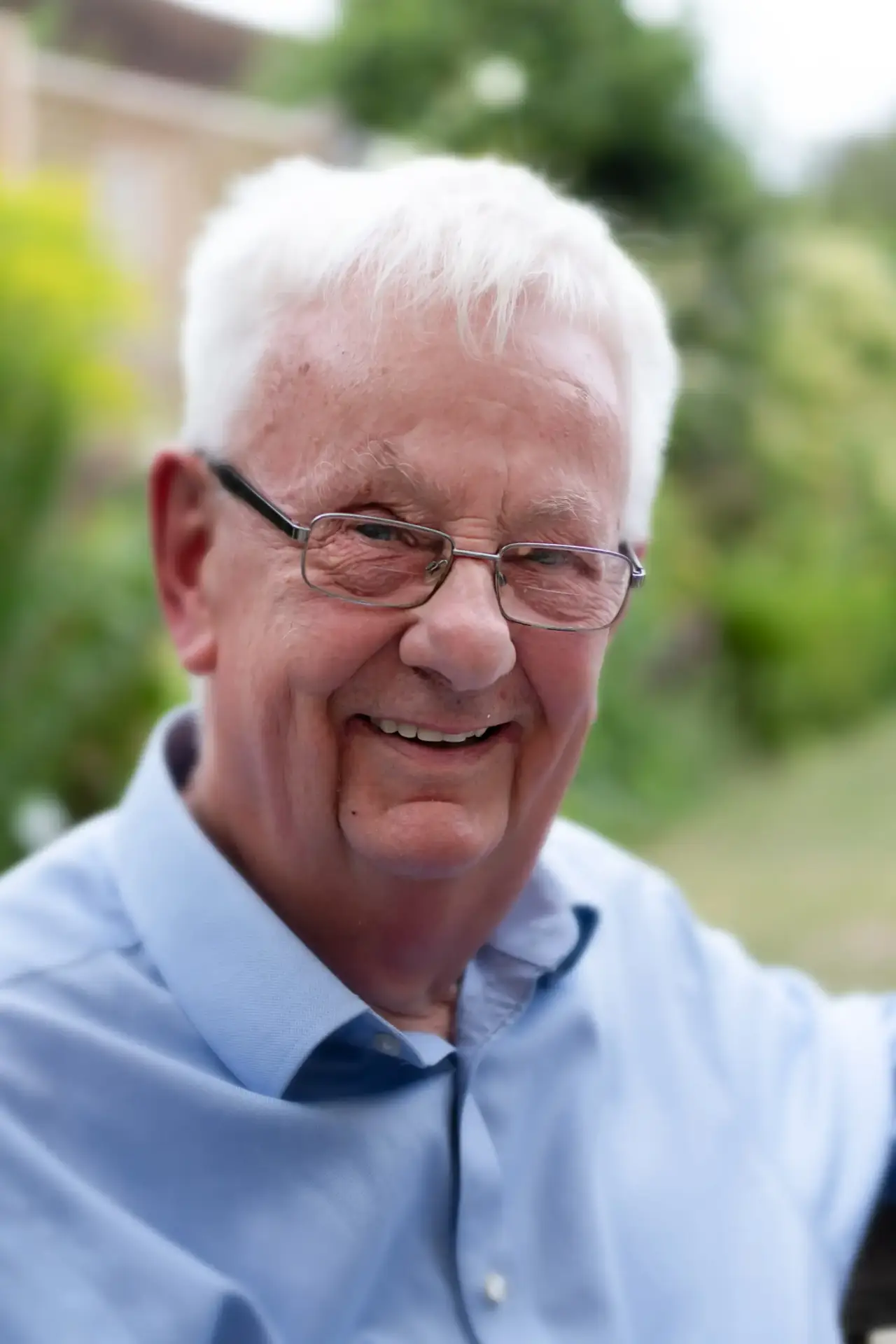 lderly man with glasses smiling outdoors in a light blue shirt.