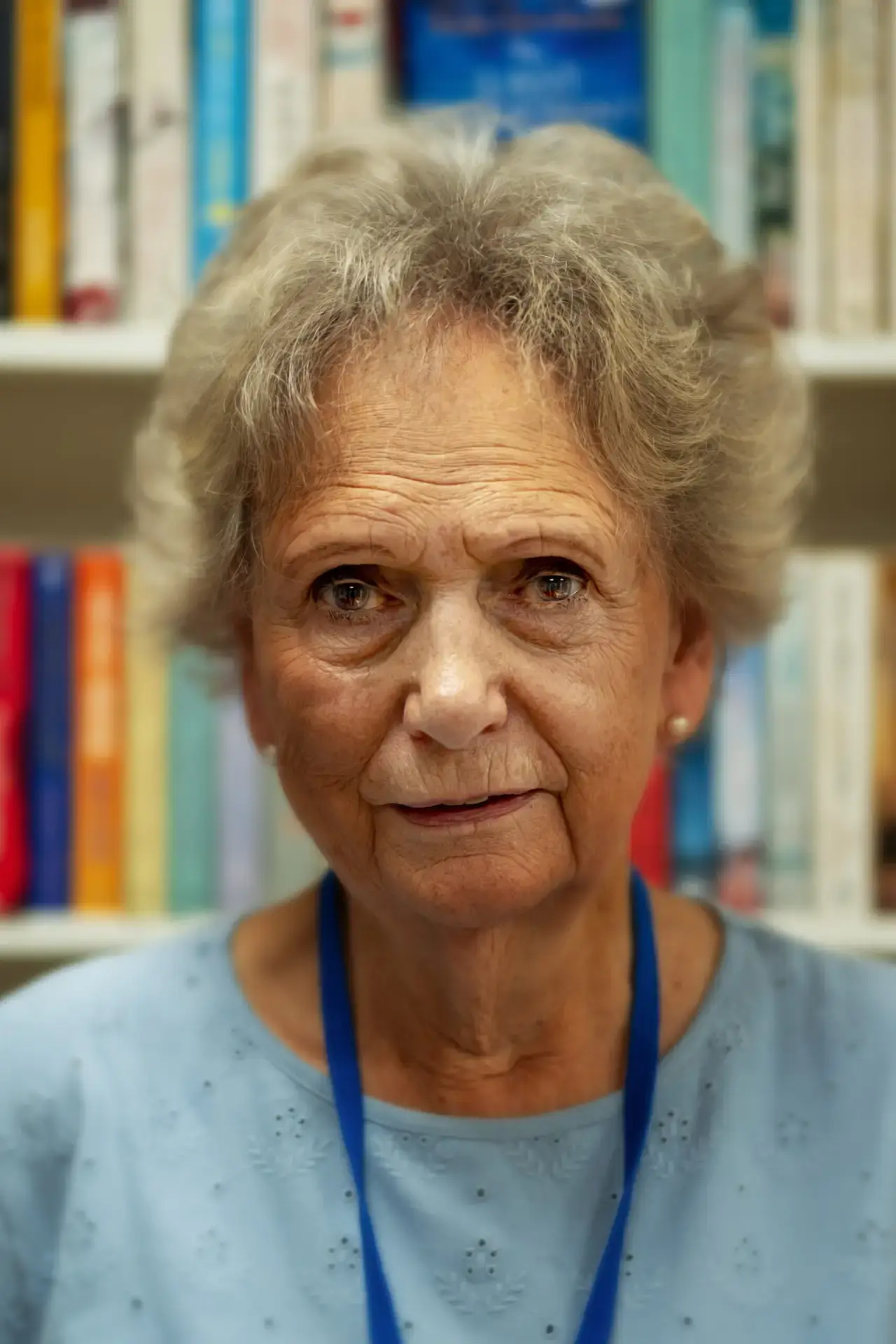 Older woman with short grey hair wearing a blue top and lanyard in front of bookshelves