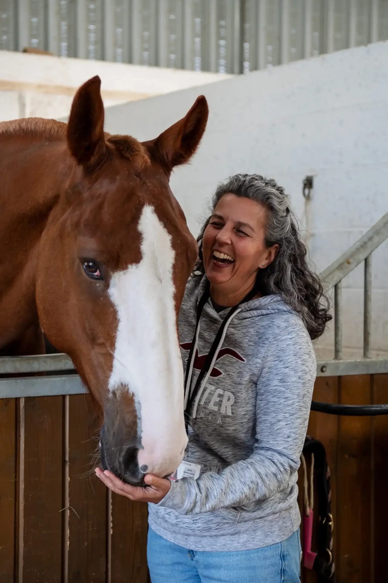 Smiling woman in grey hoodie feeding and petting a brown horse with a white blaze on its face
