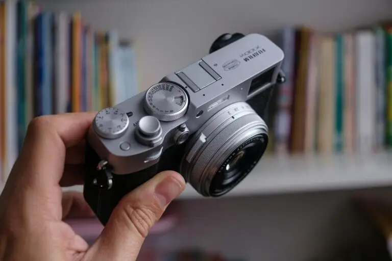 Hand holding a silver Fujifilm X100V camera with books blurred in the background