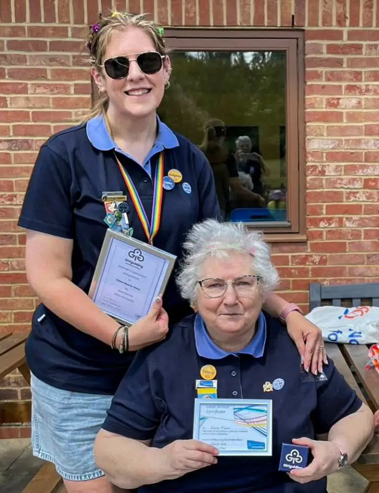 Two women in Girlguiding uniforms holding certificates and badges outside a brick building