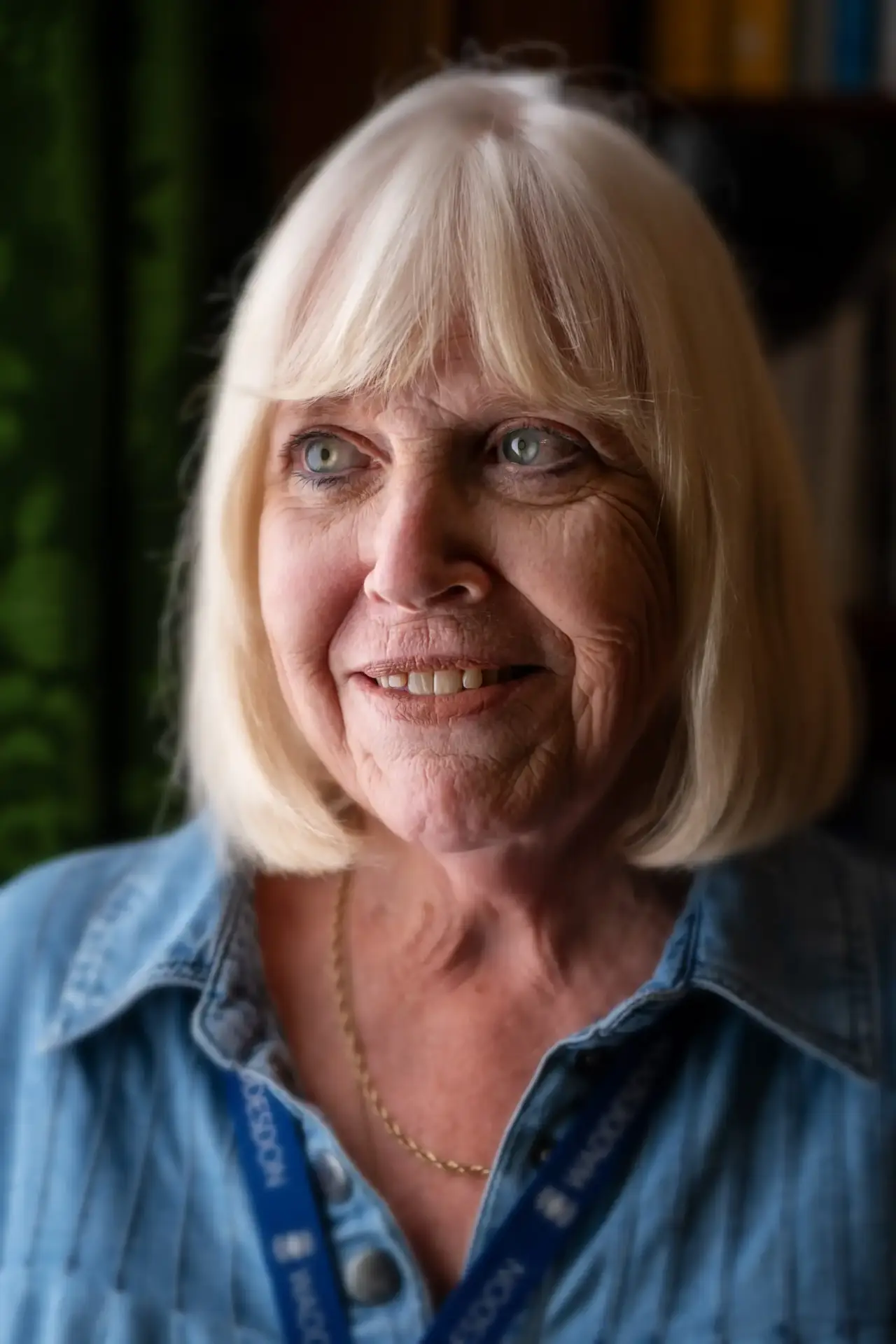 Smiling older woman with blonde hair wearing a denim shirt and lanyard indoors