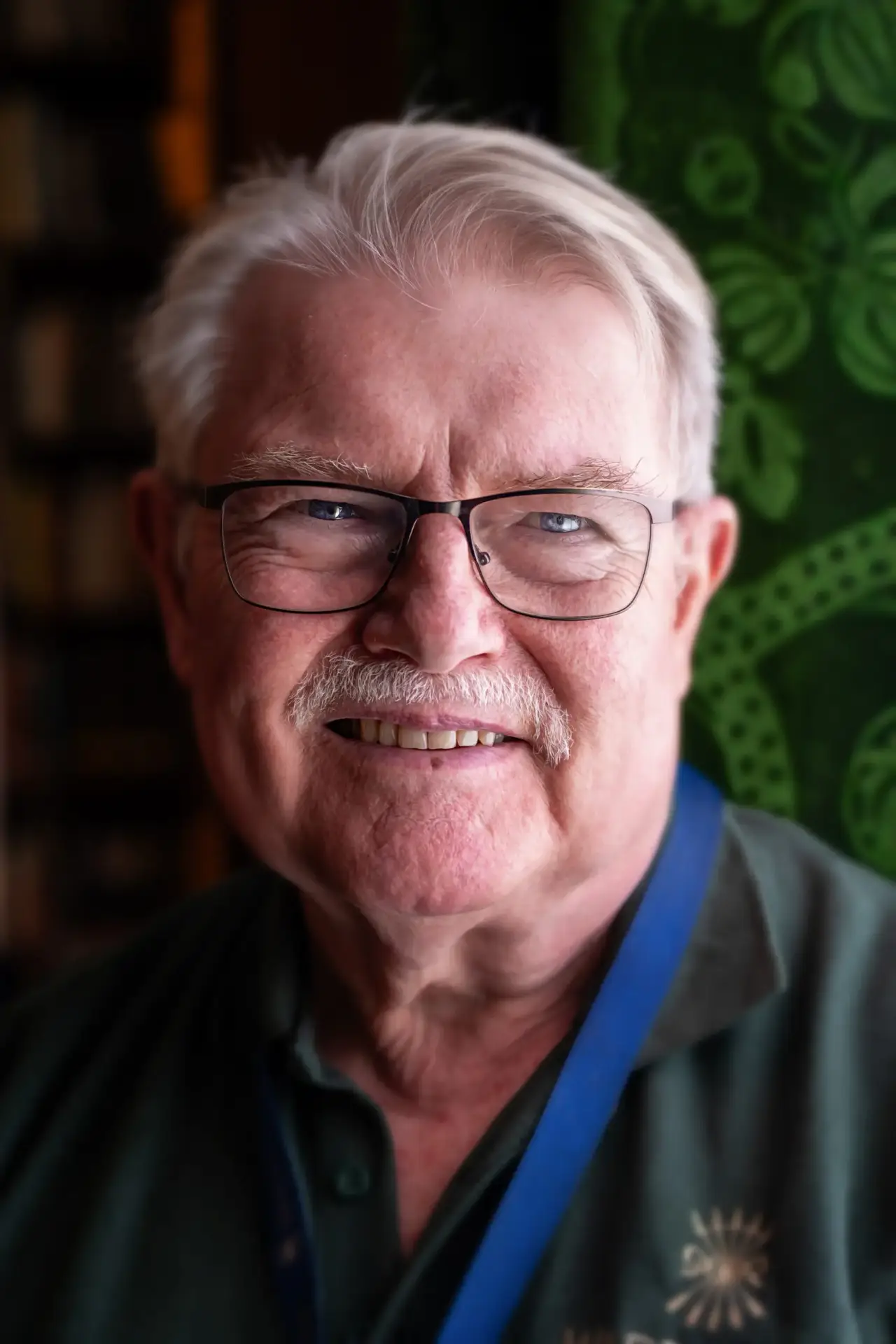 Smiling older man with glasses and grey hair wearing a green shirt and lanyard indoors