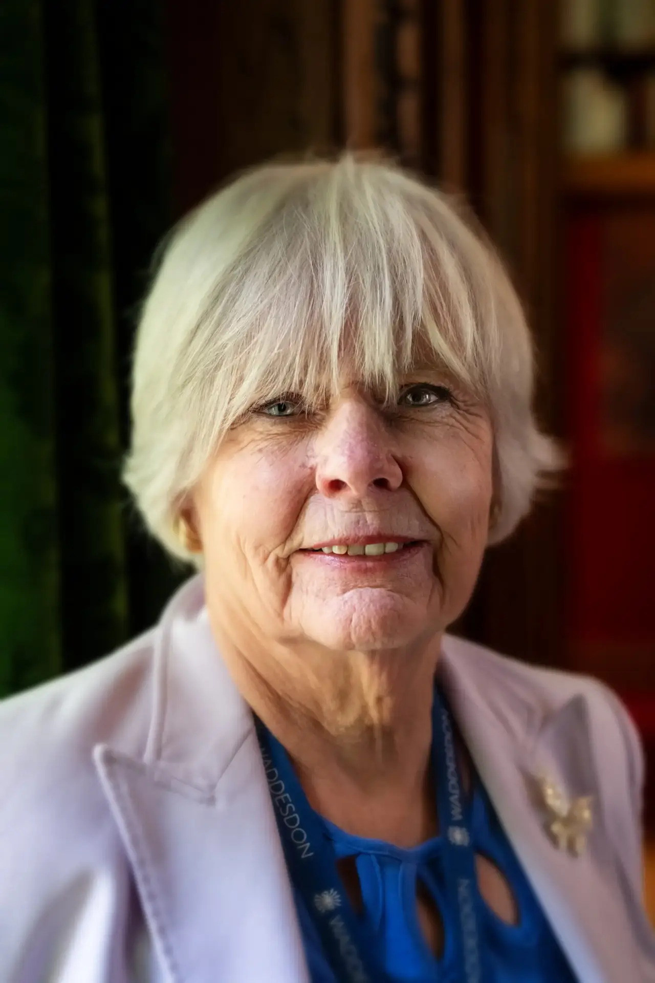 Smiling older woman with short grey hair wearing a light blazer and blue top indoors