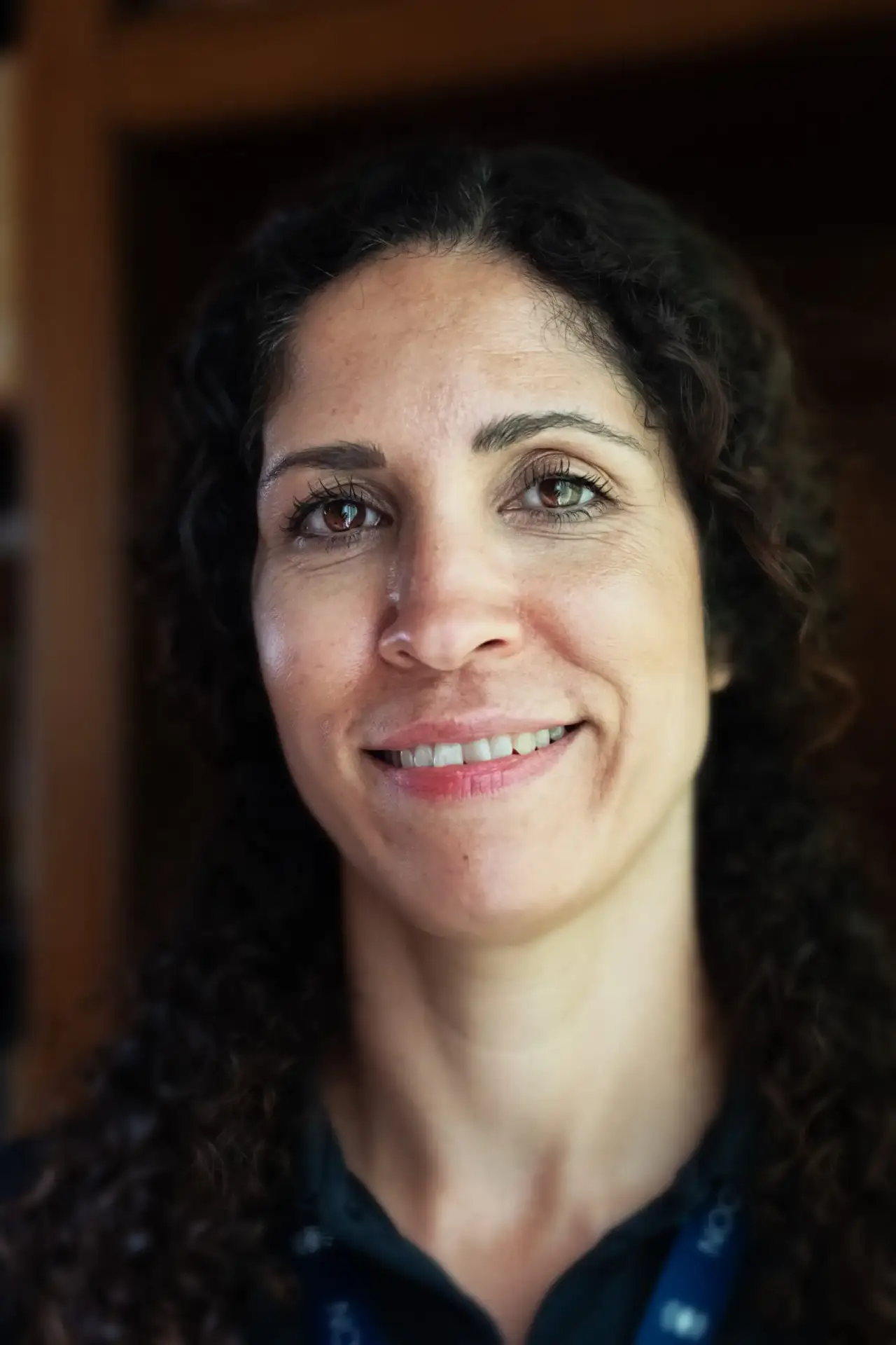 Smiling woman with curly dark hair wearing a dark shirt and lanyard indoors