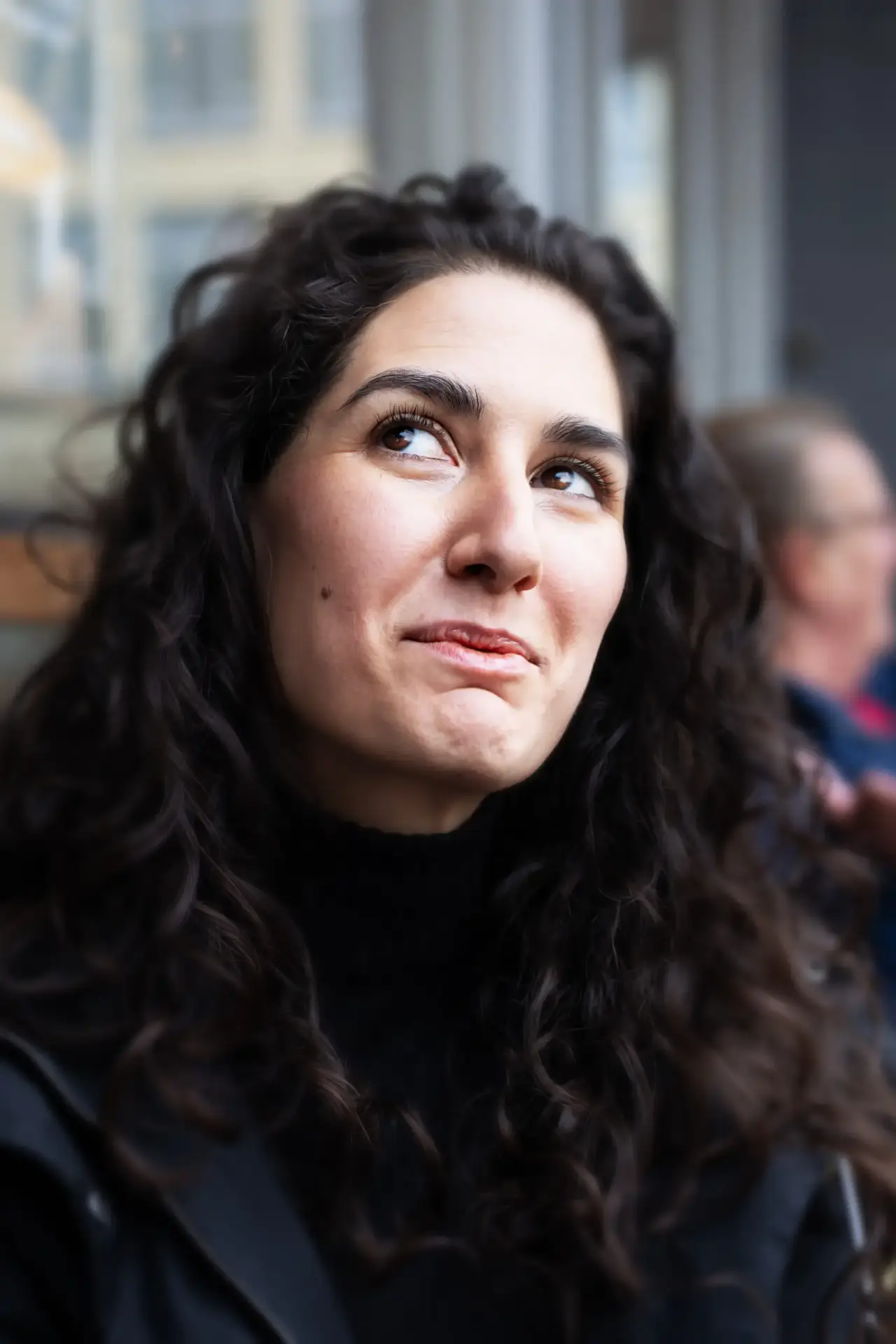 Woman with long curly dark hair looking upwards and smiling slightly while seated indoors
