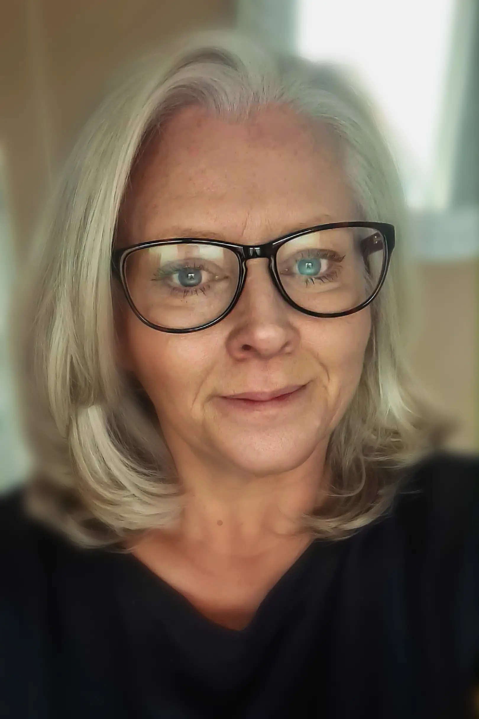 Woman with shoulder-length grey hair and glasses wearing a dark top indoors