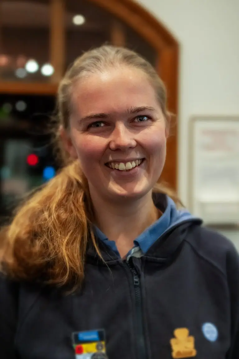 Smiling young woman with blonde hair tied back wearing a navy hoodie and badges indoors