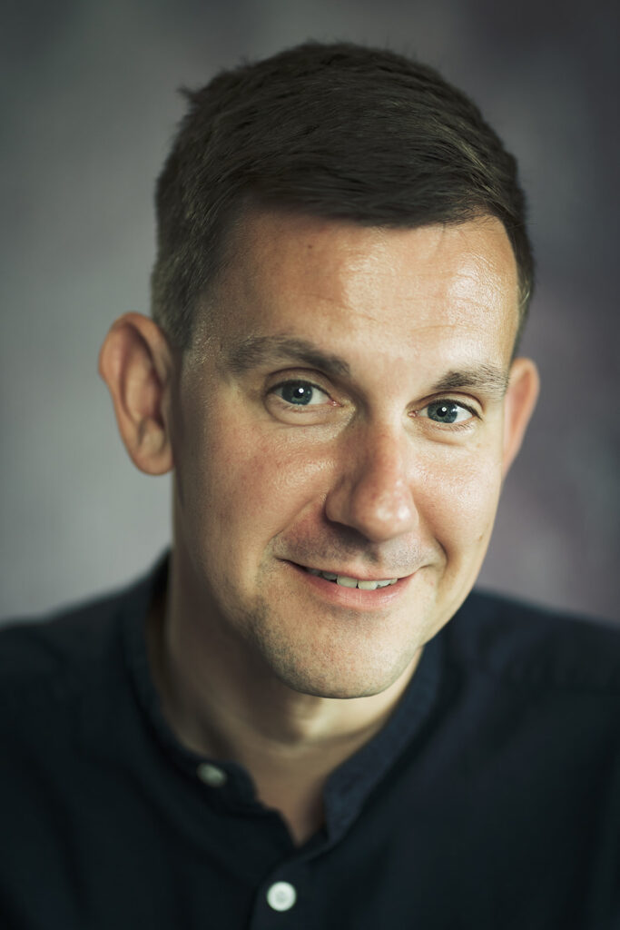 Smiling male actor in a black shirt photographed with soft light in St Albans, Hertfordshire.