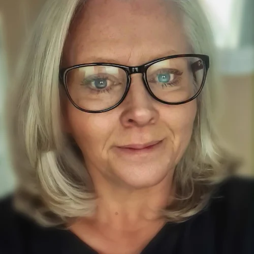 Woman with shoulder-length grey hair and glasses wearing a dark top indoors