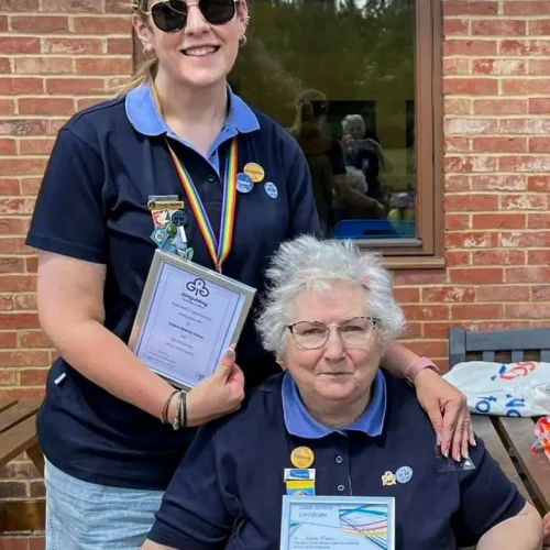 Two women in Girlguiding uniforms holding certificates and badges outside a brick building