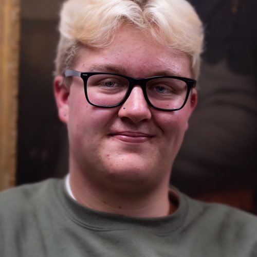 Young person with blonde hair, glasses, and nose stud wearing a green sweatshirt