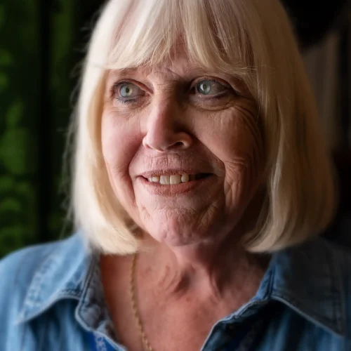Smiling older woman with blonde hair wearing a denim shirt and lanyard indoors