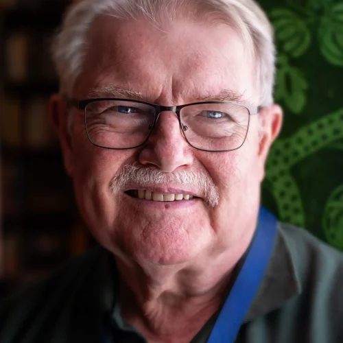 Smiling older man with glasses and grey hair wearing a green shirt and lanyard indoors