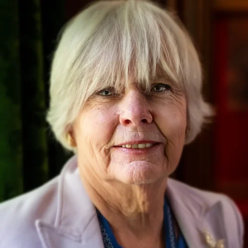 Smiling older woman with short grey hair wearing a light blazer and blue top indoors