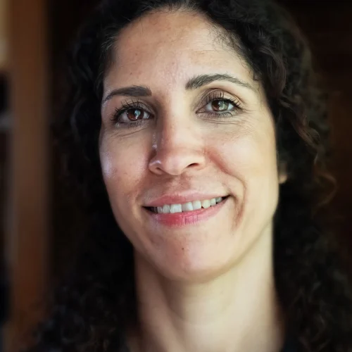 Smiling woman with curly dark hair wearing a dark shirt and lanyard indoors