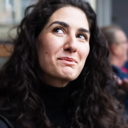 Woman with long curly dark hair looking upwards and smiling slightly while seated indoors