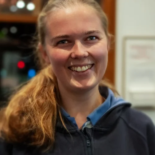 Smiling young woman with blonde hair tied back wearing a navy hoodie and badges indoors