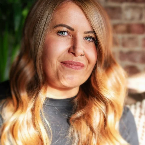 Smiling woman with long wavy blonde hair and a nose ring wearing a black top indoors